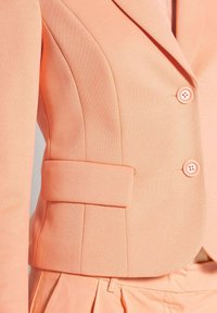 Perzikkleurige blazer met een gestructureerde stof, een getailleerd ontwerp, schoudervullingen, twee ronde knopen en een zijzak voor praktisch gebruik.