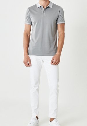 Mand iført lysegrå kortærmet poloshirt med hvid kant, hvide bukser og hvide sneakers, der står mod en ensfarvet baggrund.