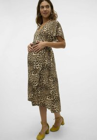 Obleka za nosečnice z leopardjim vzorcem, kratkimi rokavi in V-izrezom, s prosto silhueto. Rumeni čevlji z nizko peto in trakovi dopolnjujejo obleko.