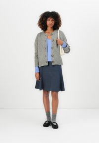 Woman with curly hair wearing gray buttoned cardigan over blue top, navy skirt, gray socks, black shoes, and holding a white shoulder bag.