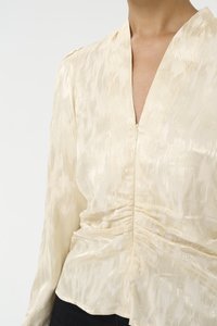 Light beige blouse with a textured pattern, v-neck, long sleeves, and gathered details at the waist. Smooth fabric with subtle sheen.