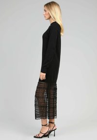 Un pull noir à coupe décontractée associé à une jupe en dentelle noire avec des motifs géométriques, portée avec des sandales à talons hauts à lanières.