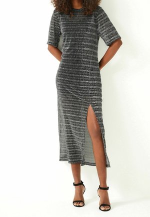 Frau trägt ein grau geripptes Midikleid mit ellbogenlangen Ärmeln und einem Oberschenkelschlitz, kombiniert mit schwarzen Sandaletten mit Knöchelriemen.