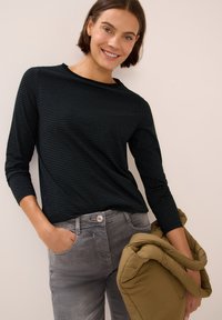 Zwarte en donkergroene gestreepte langemouwen top gedragen met grijze jeans. Het model houdt een opgevouwen olijfgroene pufferjack vast.
