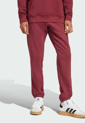 Burgundy jogger pants made of soft fabric, featuring an elastic waistband and cuffs, paired with white sneakers having black stripes.