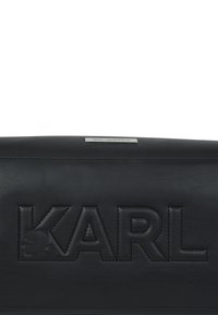 Μαύρη δερμάτινη επιφάνεια με ανάγλυφο μεγάλο κείμενο "KARL" και μια μικρή μεταλλική πλάκα από πάνω με "KARL LAGERFELD" χαραγμένο.