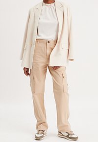 Beige bomulds cargo bukser med sidelommer, parret med en cremet bluse med ribbet tekstur og en skræddersyet cremet blazer. Sneakers inkluderet.