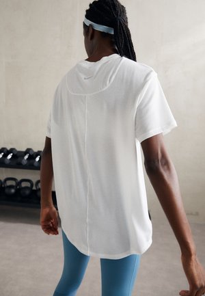 Persoon met gevlochten haar, gekleed in een wit shirt, blauwe legging en een wit hoofdband, staande in een sportschool met gewichten en kettlebells op de achtergrond.