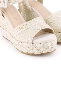 Sandalias de cuña con correas de color beige trenzadas, detalle de trenzado y plantilla acolchada. Presenta un pequeño emblema dorado en la correa.