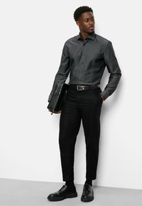 Donkergrijze overhemd met textuur, zwarte dresspantalons en zwarte glanzende schoenen. Houdt een zwarte leren aktetas vast.