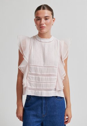 Kvinne med tilbakelent hår iført en hvit ermeløs bluse med volanger på skuldrene og blonde detaljer, kombinert med blå høytlivsskjørt jeans.