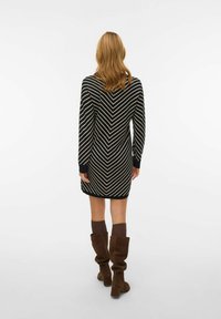 Juodos ir kreminės spalvos chevrono dryžuota megzta suknelė su ilgomis rankovėmis, prigludusia siluetu ir ribotais rankogaliais, derinama su ruda iki kelių siekiančiomis aulinėmis.