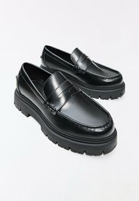 Paire de mocassins en cuir noir avec semelles épaisses en caoutchouc à crampons, détail de bride penny et bouts arrondis sur fond clair.