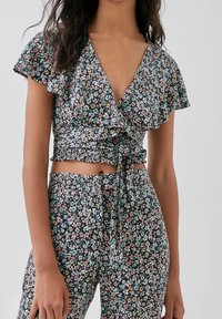 Haut court cache-cœur à fleurs avec fond noir, manches courtes à volants et ourlet volanté. Présente de petites fleurs multicolores et un détail noué.