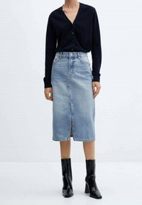 Jupe midi en denim bleu clair avec une fente sur le devant, associée à un cardigan noir ajusté. Complétée par des bottines à talons noirs.