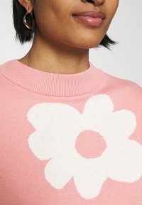 Ljust rosa tröja med en stor vit blomstergrafik. Har en ribbad halsringning och mjukt, texturerat tyg.