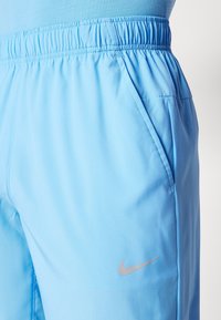 Lichtblauwe Nike sportbroek met een elastische tailleband, zijzakken en een klein zilveren logo op het linkerbeen. Gladde textuur.