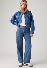 Denimjack en hooggekruiste blauwe spijkerbroek, beide in een gemiddelde wastoe. De jack heeft knoopsluitingen, twee borstzakken en een ontspannen pasvorm.