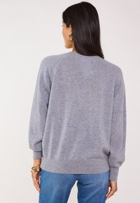 Donna con lunghi capelli scuri che indossa un morbido maglione di un leggero viola e jeans blu, vista di spalle con orecchini ad anello dorati e bracciali.