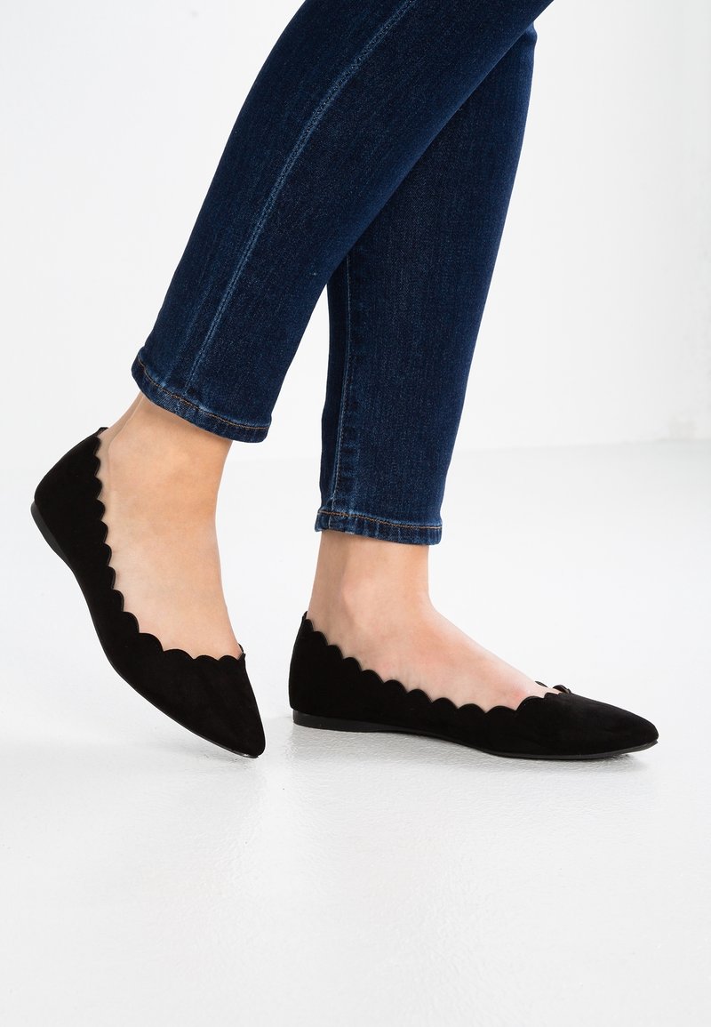 Pies calzando planos de ante negro con borde en forma de ola y punta, combinados con jeans ajustados de color azul oscuro, sobre un fondo blanco.