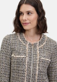 Marynarka typu tweed w kolorach czarnym, białym i kremowym, z frędzlami, zamkiem błyskawicznym i dwiema naszytymi kieszeniami. Strukturalny fason z okrągłym dekoltem.