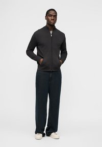 Άνδρας που στέκεται με τα χέρια στις τσέπες, φοράει μαύρο μπουφάν με φερμουάρ, σκούρο φαρδύ παντελόνι και λευκά αθλητικά παπούτσια μπροστά από απλό φόντο.