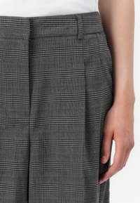 Gros plan d'une personne portant un pantalon à carreaux gris avec des passants de ceinture et des plis, et un haut blanc visible à la taille.