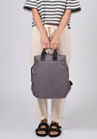 Mochila gris con material acolchado, forma cuadrada, asas superiores y cremallera lateral. El modelo lleva puesta un pantalón ligero y sandalias.