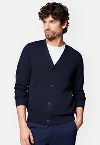 Ciemnogranatowy kardigan, wykonany z lekkiego dzianiny, z dekoltem w kształcie V, sześcioma guzikami z przodu, ściągaczami na mankietach i u dołu, noszony na białej koszuli.