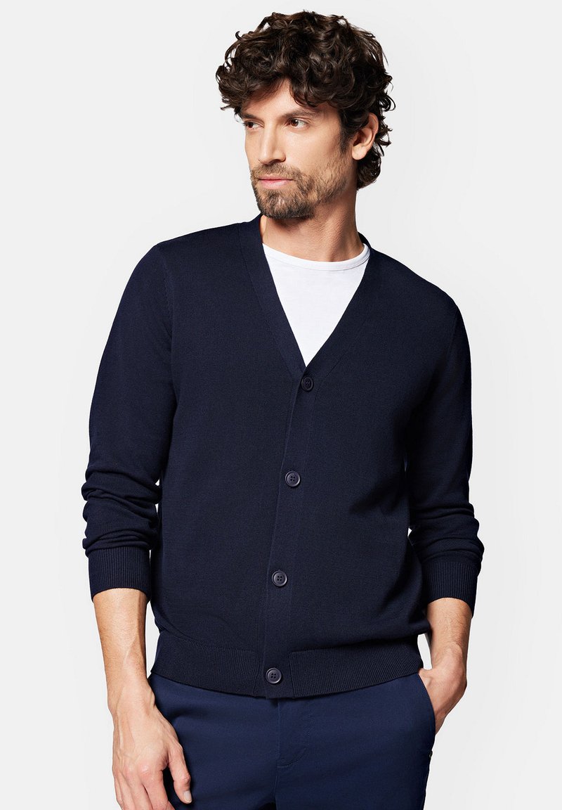 Ciemnogranatowy kardigan, wykonany z lekkiego dzianiny, z dekoltem w kształcie V, sześcioma guzikami z przodu, ściągaczami na mankietach i u dołu, noszony na białej koszuli.