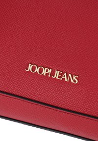 Borsa in pelle sintetica rossa con texture, caratterizzata da un logo dorato "JOOP! JEANS". Linea nera liscia vicino alla parte inferiore.