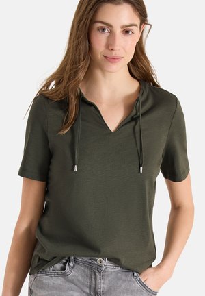 Donna che indossa una maglietta verde oliva a maniche corte con scollo a V e coulisse, abbinata a jeans grigio chiaro, in piedi con una mano in tasca.