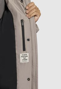 Gai bēšas krāsas polsterēta jaka ar melnu oderi, rāvējslēdzēja kabatu un izšūtu etiķeti ar tekstu "Siltas apskāviens izgatavots no auduma." Mīksta tekstūra.