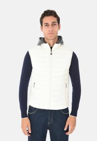 Uomo che indossa un gilet senza maniche bianco trapuntato sopra una camicia a maniche lunghe blu navy e jeans scuri, in piedi contro uno sfondo chiaro e uniforme.