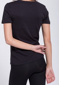 Camiseta negra de manga corta, hecha de algodón suave, con un escote redondeado y un diseño ajustado, mostrada desde la espalda con un brazo apoyado en la cadera.