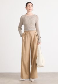 Beige, gebreide top met lange mouwen met een textuurontwerp, gecombineerd met hooggesneden, geplooide, lichtbruine wijde broek. Houdt een crèmekleurige handtas vast.