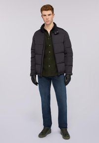 Svart vadderad jacka med hög krage, bärs över en mörkgrön skjorta, i kombination med blå jeans och mörkgröna sneakers. Handskarna är i svart läder.