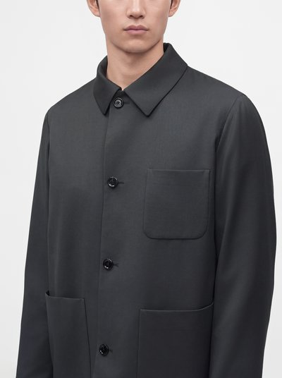 Homme portant un manteau boutonné gris foncé avec un col et trois grandes poches avant, sur un fond clair uni.