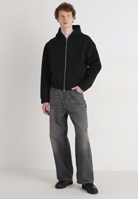 Sweat à fermeture éclair noir avec une poche kangourou, porté avec un jean large gris délavé et des chaussures noires. Tissu lisse et coupe décontractée.