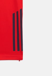 Κόκκινη αθλητική φανέλα με ναυτικό μπλε τριπλή γραμμή σχεδίου στους ώμους. Φτιαγμένη από λείο, ελαφρύ ύφασμα με εφαρμοστό σχέδιο.