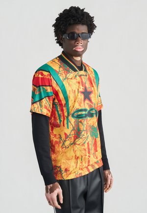 Farverig kortærmet skjorte med et levende mønster i gult, rødt og grønt. Har en krave og en sort langærmet undertrøje nedenunder.