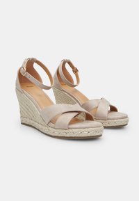 Beige wedge sandals with crossed straps and adjustable ankle buckle, featuring a rope-woven jute sole and smooth suede upper.