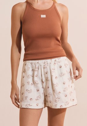 Vrouw draagt een bruin geribd tanktop en witte shorts met elastische taille met koffie- en croissantprints op een beige achtergrond.