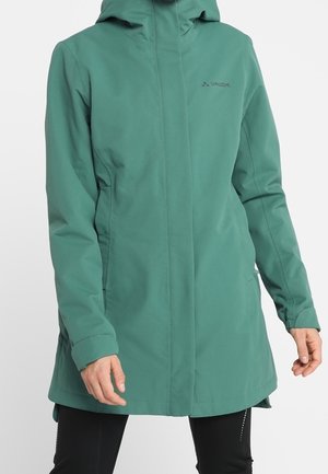 Vrouw draagt een groen knielang gewatteerd jack met capuchon, lange mouwen en een ritssluiting aan de voorkant, gecombineerd met een zwarte broek.