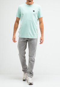 Mint green short-sleeve t-shirt with a round neck and small black logo on the chest, paired with light gray jeans and multicolored sneakers.