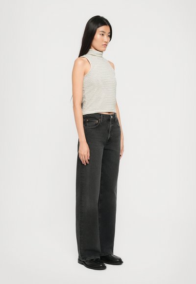 Vrouw met lang zwart haar staat in een mouwloze witte coltrui, high-waisted zwarte wijde jeans en zwarte schoenen op een witte achtergrond.