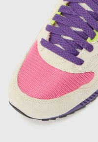 Κοντινή λήψη της μύτης ενός αθλητικού παπουτσιού με ροζ δίχτυ, μπεζ σουέτ περίγραμμα, μωβ κορδόνια και πράσινα μεταλλικά πατήματα κορδονιών σε λευκό φόντο.