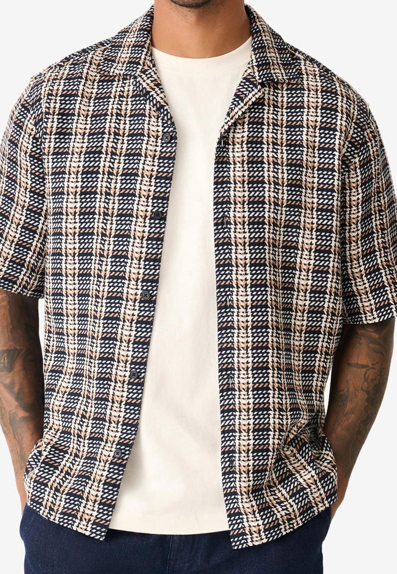 Homme portant une chemise à manches courtes boutonnée avec un motif pied-de-poule noir, beige et blanc par-dessus un t-shirt blanc uni.