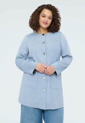 Kobieta o kręconych włosach ubrana w jasnoniebieski zapinany na guziki płaszcz i niebieskie dżinsy, stoi z rękami złożonymi na talii, delikatnie się uśmiechając.