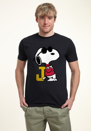 Młody mężczyzna z blond włosami, ubrany w czarną koszulkę z postacią Snoopy’ego w okularach przeciwsłonecznych i czerwony sweter, stoi z rękami w kieszeniach.